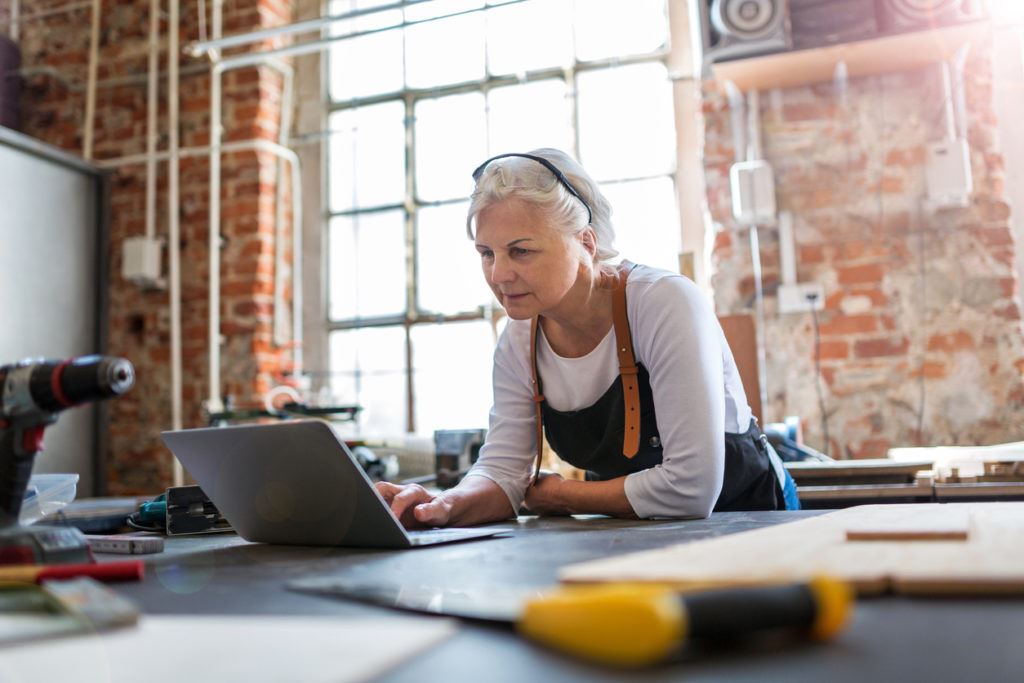 An older woman, in a workshop with a large window behind her, looking intently at a laptop screen.